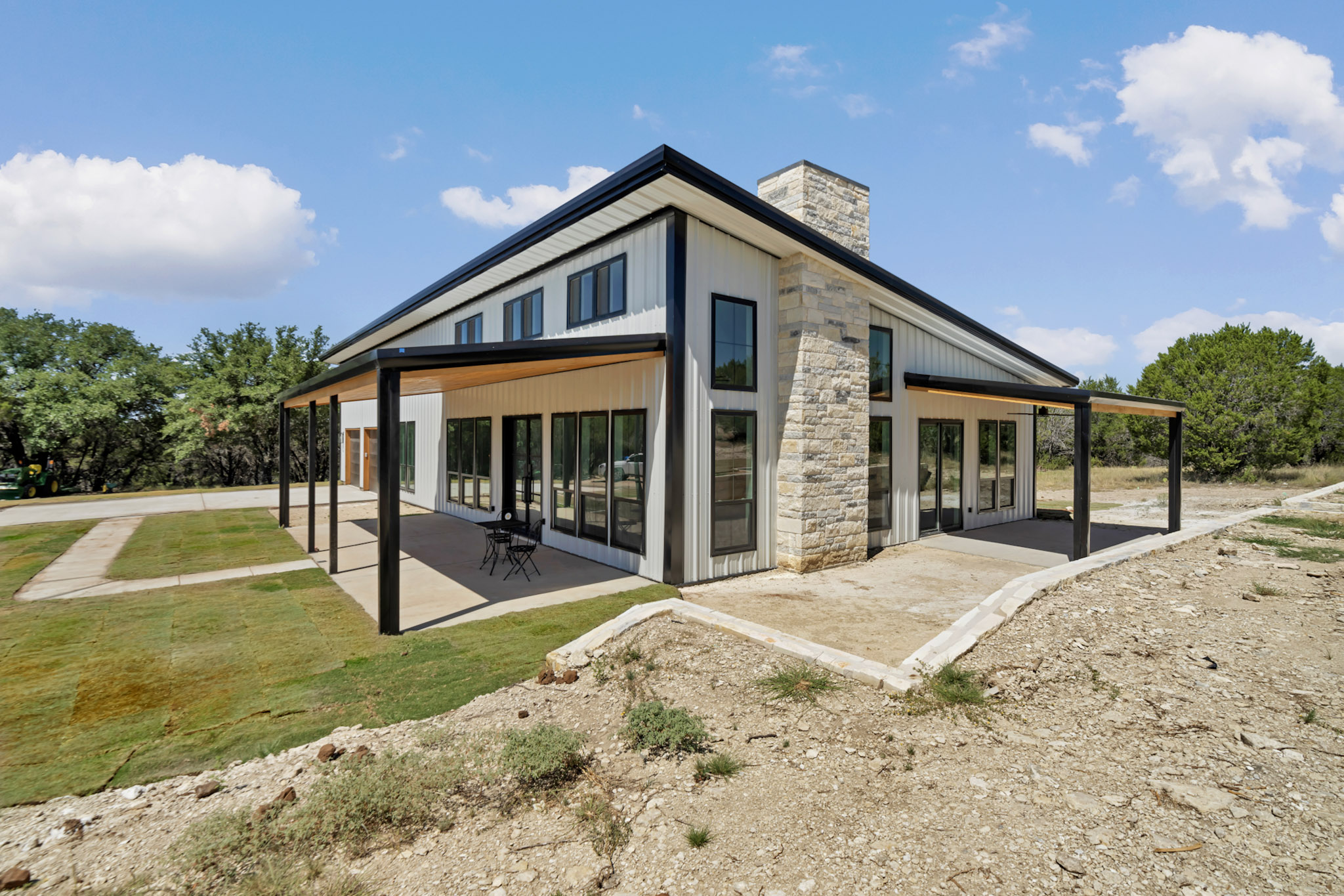 White metal barndominium with black trim, a stone chimney and a wraparound covered porch