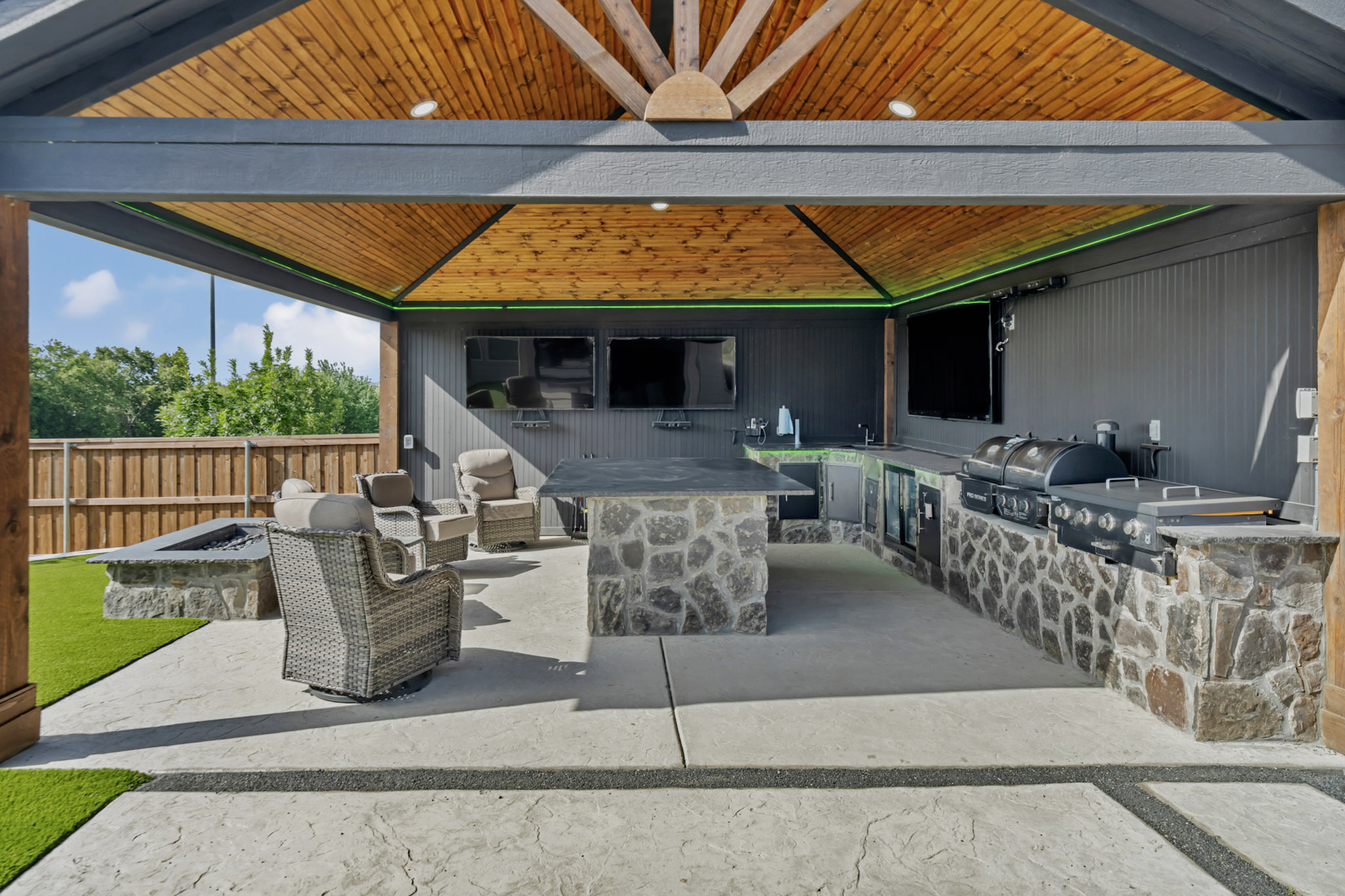 Covered outdoor kitchen with a stone island, built in grills, mounted TVs and a gas fire pit